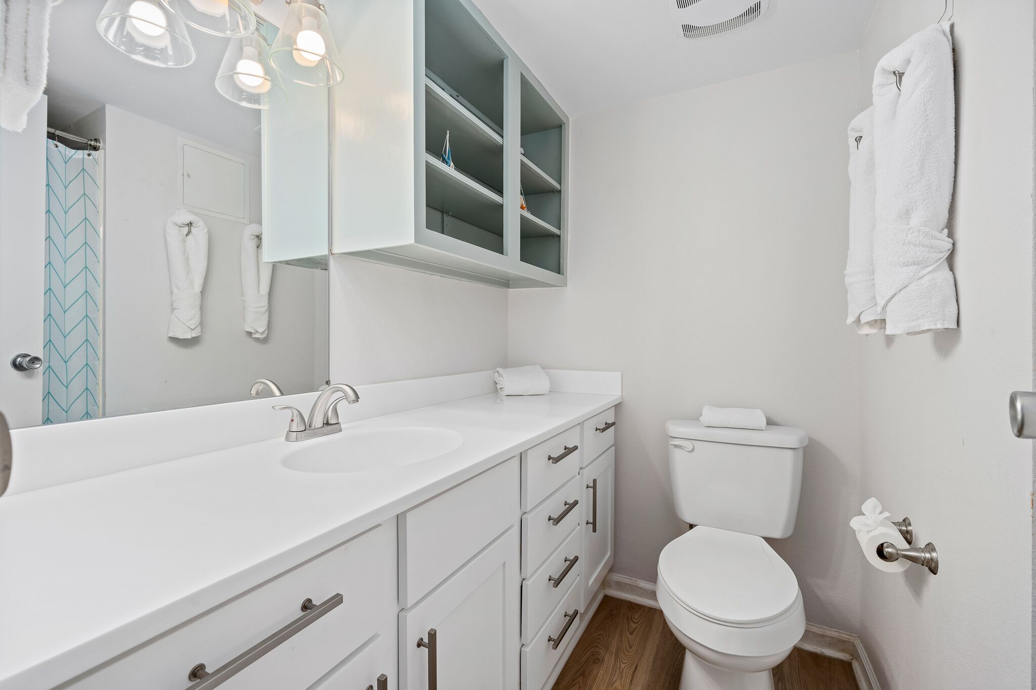 Une salle de bain moderne avec un lavabo, un miroir et des toilettes, le tout de couleur blanche. Des serviettes blanches sont accrochées au mur, et un rideau de douche orné d’un motif bleu est visible.