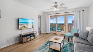 Ein Wohnzimmer mit einem Flachbildfernseher, einem Deckenventilator, einem Holztisch, einem grauen Sofa und Meerblick durch eine Schiebetür.