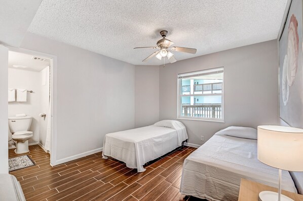 A bedroom with two single beds, a ceiling fan, a window with a balcony view, and a white toilet in the adjacent bathroom.