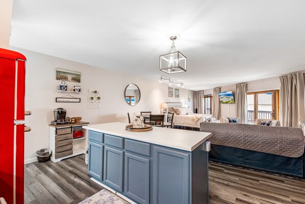 A compact hotel room with a kitchenette, a bed with a quilted cover, a dining area with a table and chairs, a red door, and a large window with curtains. - La Mirage Condo 240 Beachfront Resort Condominiums with Private Boardwalk to Beach (Port Aransas)