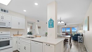 A modern kitchen with white cabinets, a dishwasher, and a microwave. There is a dining area with a table and chairs, and a view of the ocean through large windows.