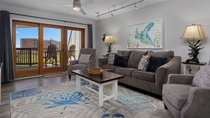 Una sala de estar con un sofá, sillones, una mesa de centro y un ventilador de techo. Hay un ventanal con vistas a edificios y un balcón.