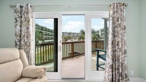 A room with a beige sofa, a sliding glass door leading to a balcony with a wooden deck and a view of a building complex.