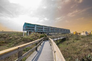 A wooden boardwalk leading to a modern building with multiple balconies, situated in a natural landscape with sparse vegetation.