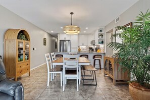 Cocina moderna con armarios blancos, un refrigerador de acero inoxidable y una zona de comedor con mesa de madera y sillas blancas.