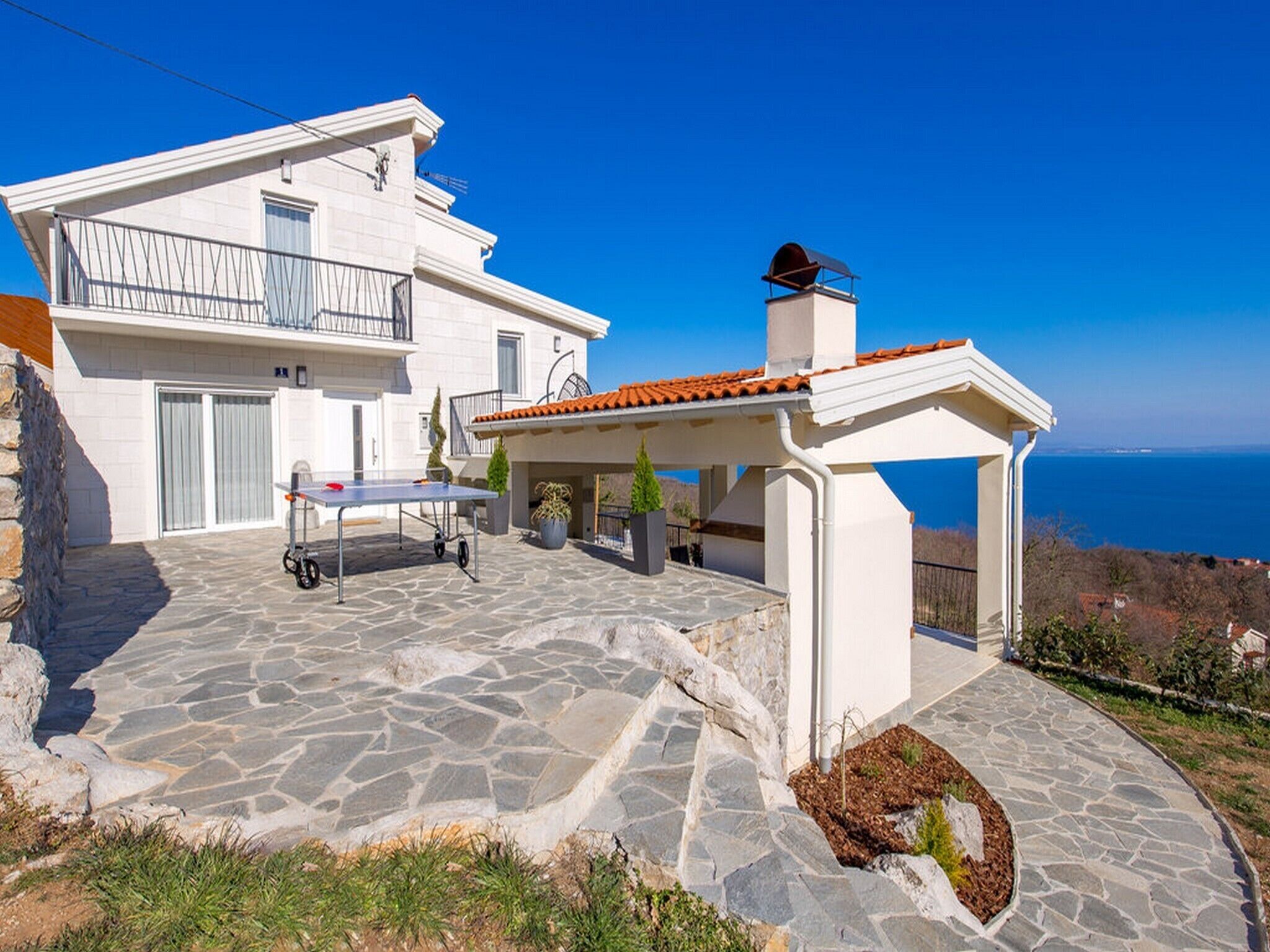 A modern white house with a stone patio, a covered outdoor seating area, and a view of the sea.