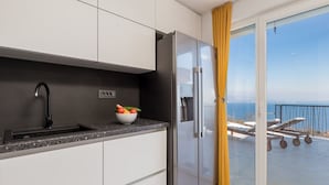 Uma cozinha moderna com uma geladeira de aço inoxidável, uma tigela de frutas no balcão e vista para o mar através de uma porta de vidro deslizante.