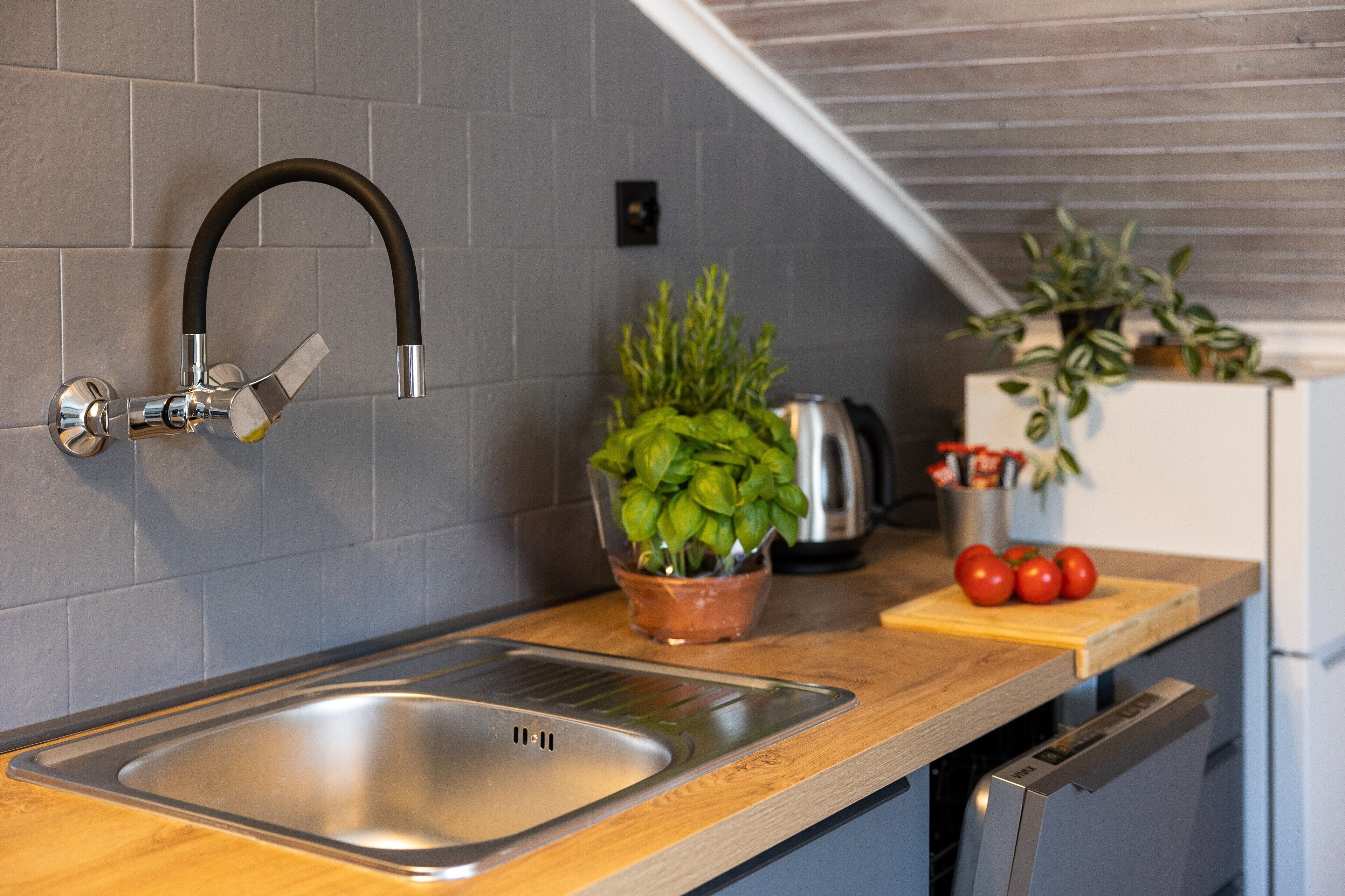 A modern kitchen with a stainless steel sink, a curved faucet, a wooden countertop, a potted basil plant, a cutting board with tomatoes, and a kettle.