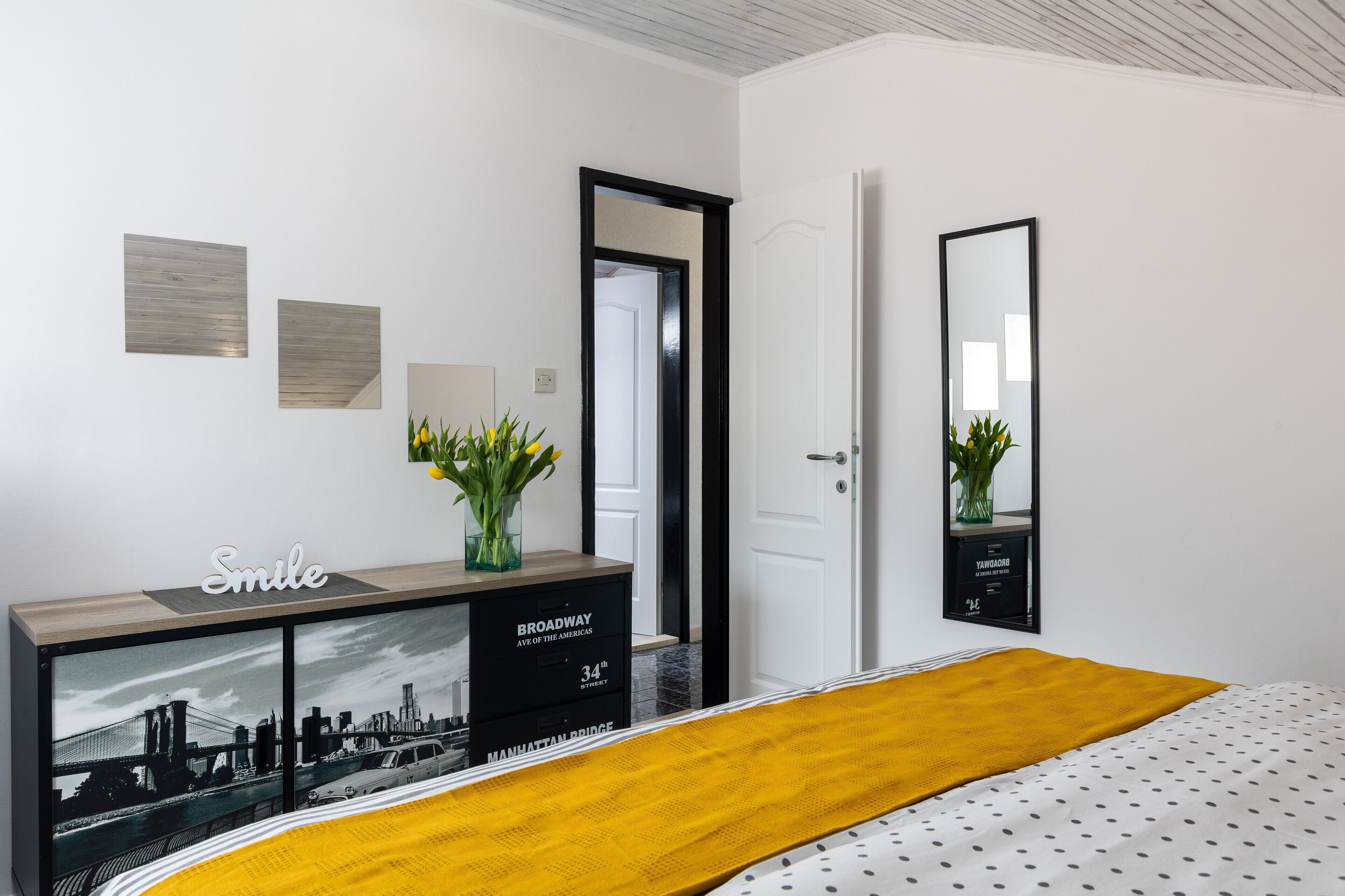 A bedroom with a bed, a bedside table with a mirror, a dresser, and a wall with a cityscape picture.