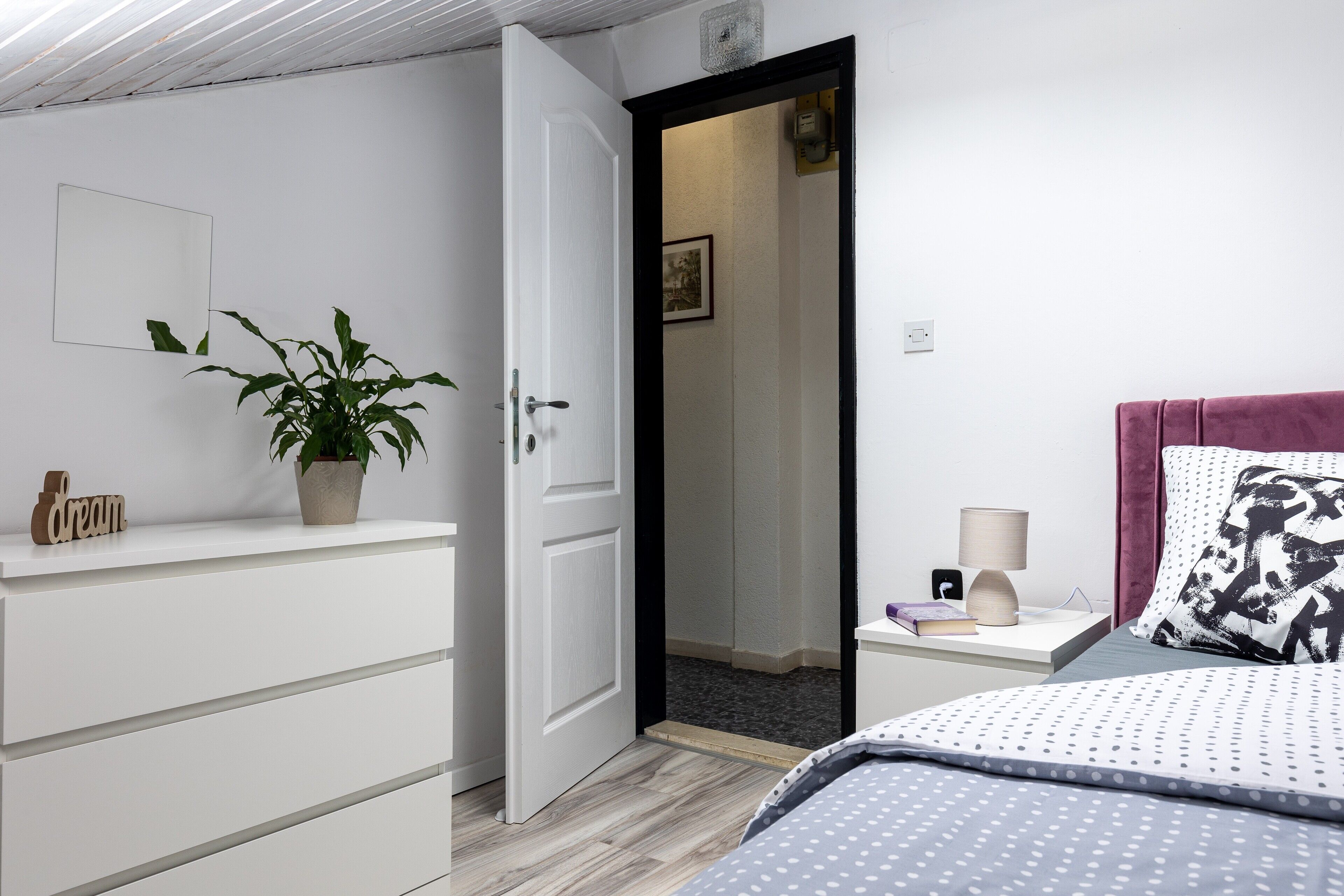 A bedroom with a bed, a nightstand, a potted plant, a dresser, and a door leading to another room.
