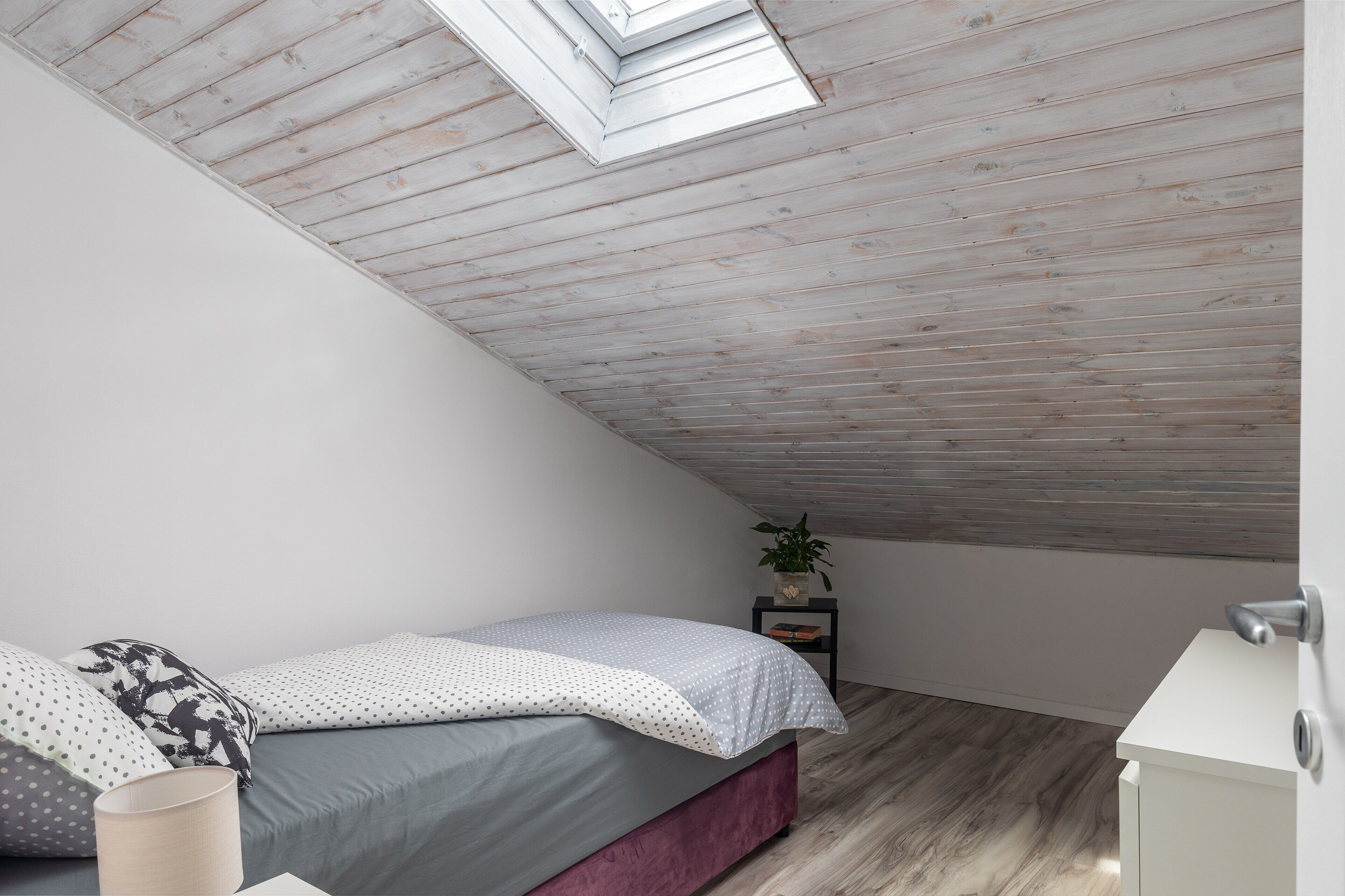A bedroom with a sloped ceiling, a skylight, a bed with grey bedding, a bedside table with a plant, and a white dresser.