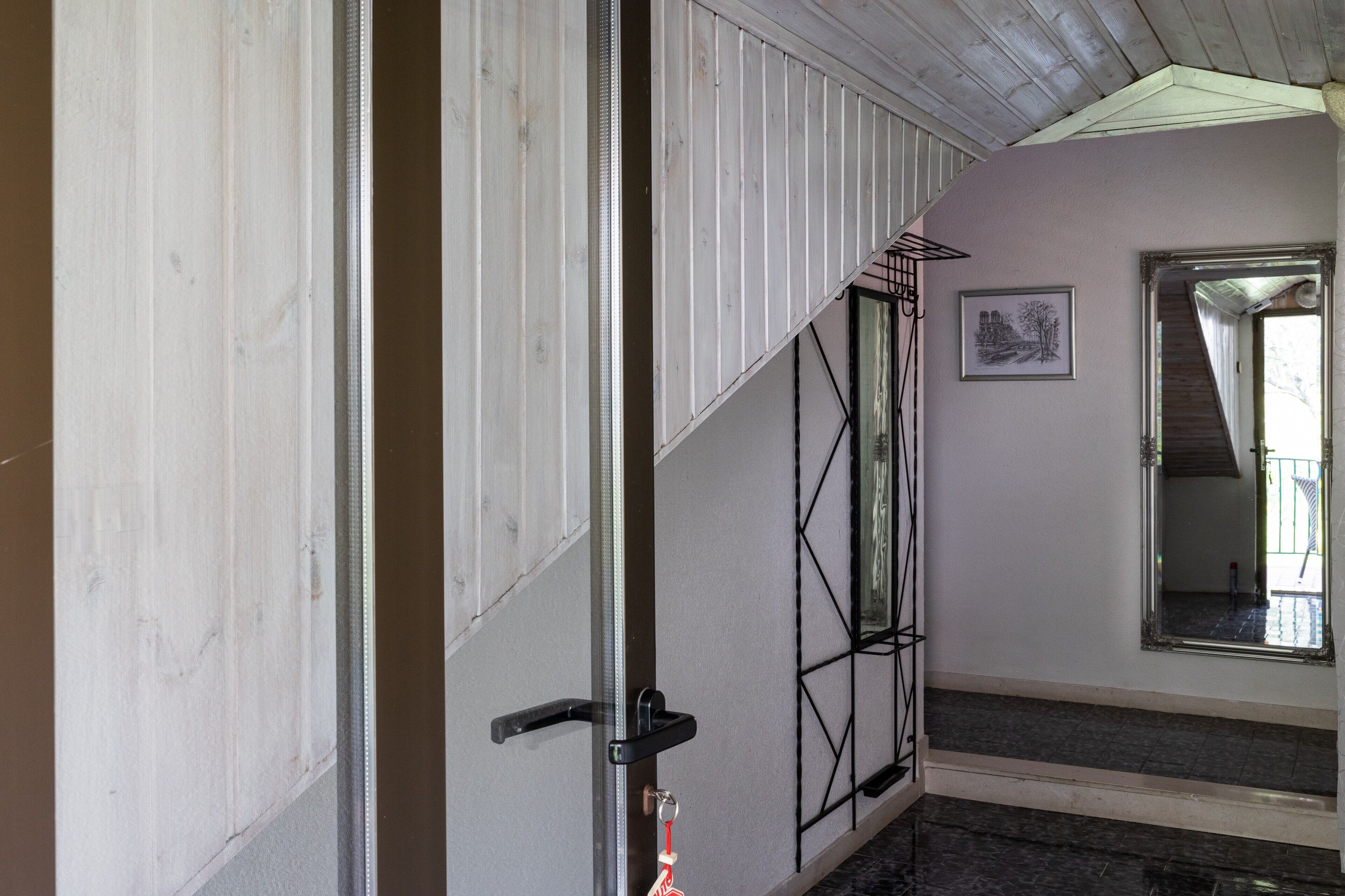 A room with a wooden ceiling, a door with a black handle, a framed picture on the wall, and a mirror reflecting a staircase.