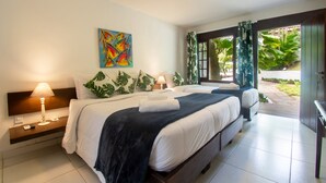 Une chambre d’hôtel avec un grand lit, une table de chevet avec une lampe, un tableau au mur et une vue sur le jardin depuis la porte.
