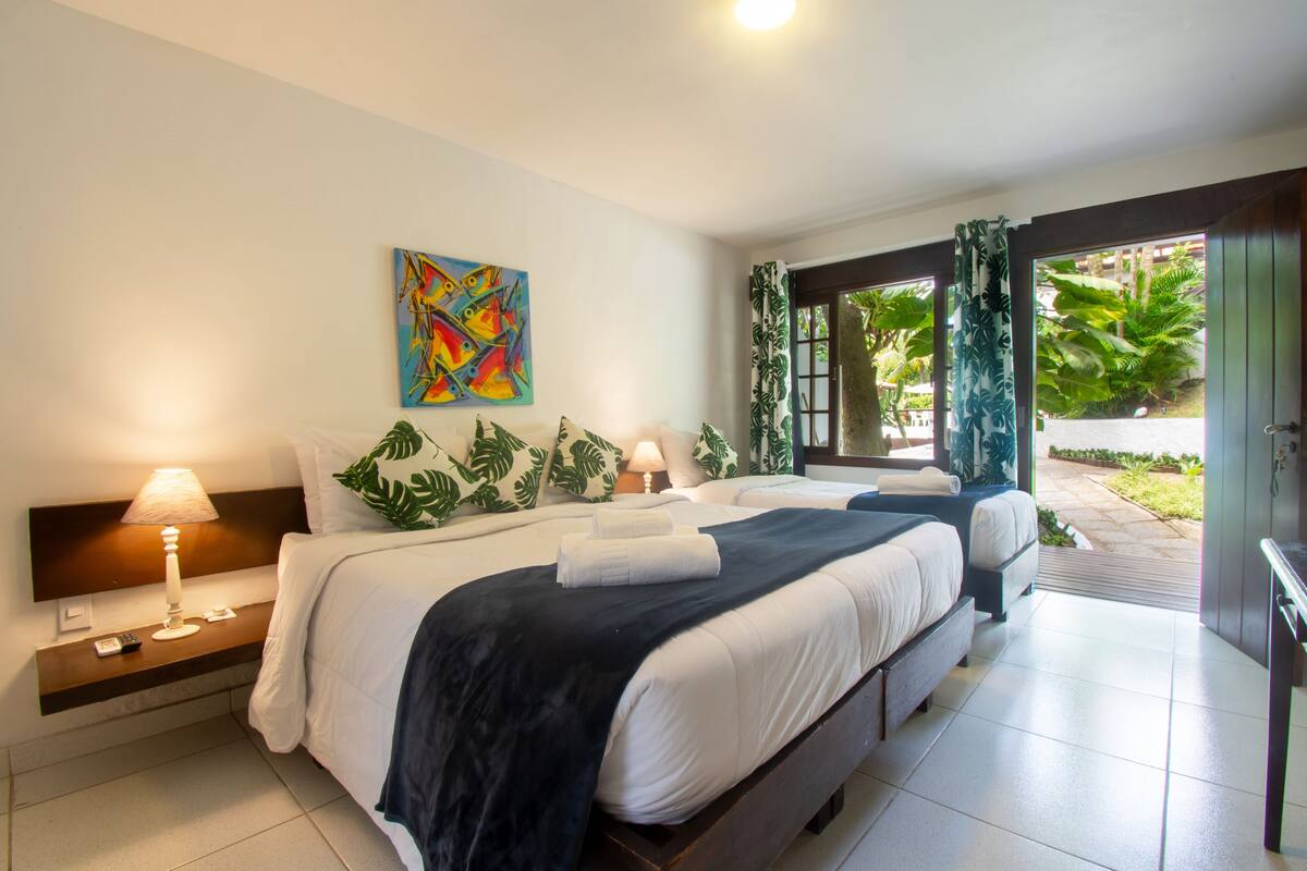 Une chambre d’hôtel avec un grand lit, une table de chevet avec une lampe, un tableau au mur et une vue sur le jardin depuis la porte.