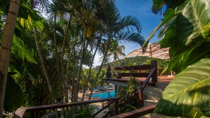 Un espace piscine entouré d’une végétation tropicale luxuriante, avec une terrasse en bois et un escalier menant à un espace salon abrité.