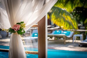 Vista de una zona junto a la piscina con sillones de descanso y una cortina blanca decorativa con flores.