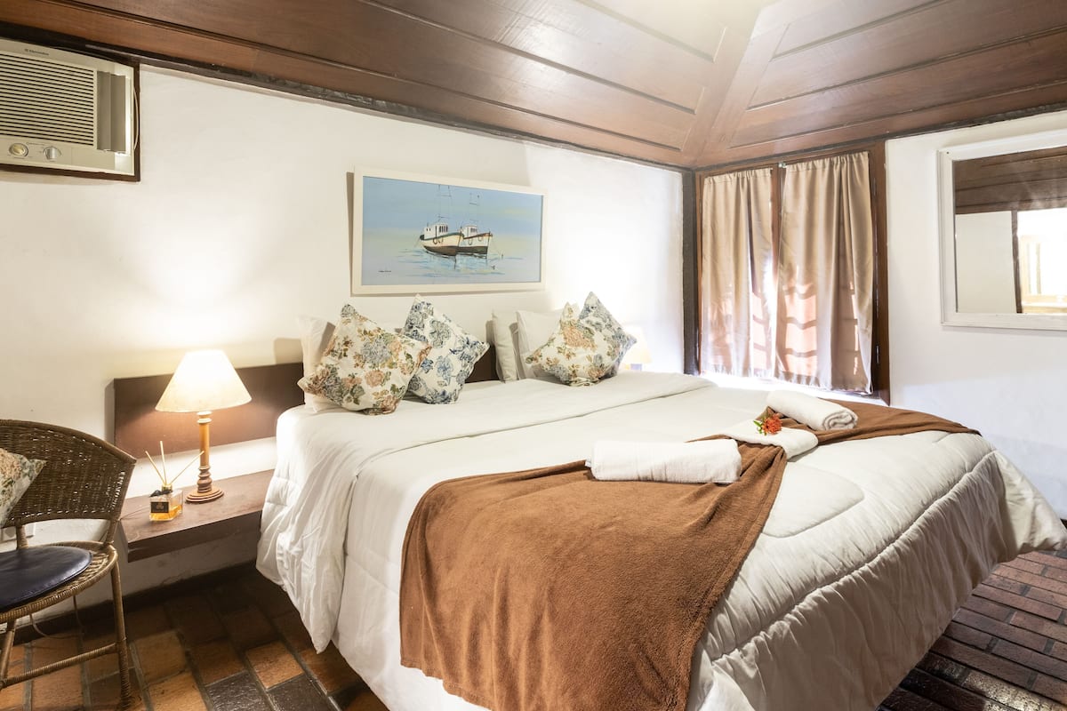 Une chambre d’hôtel avec un lit, une chaise, une lampe, un tableau et une fenêtre avec des rideaux.