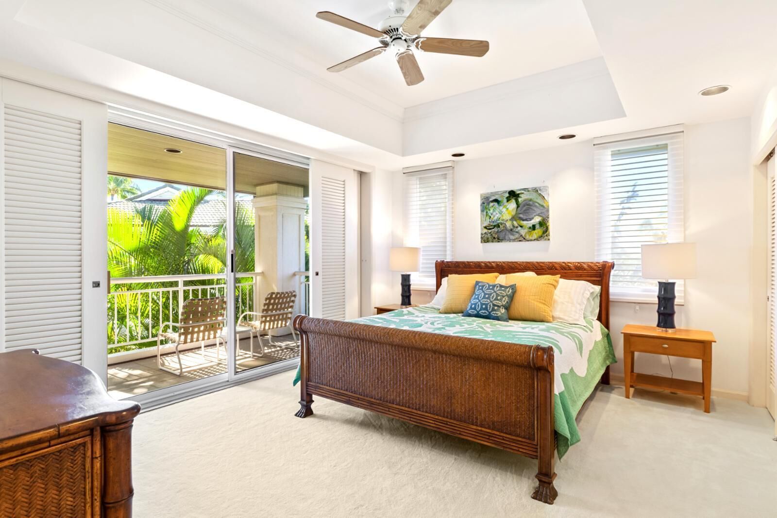 A bedroom with a large bed, a ceiling fan, a balcony with chairs, and a sliding glass door.