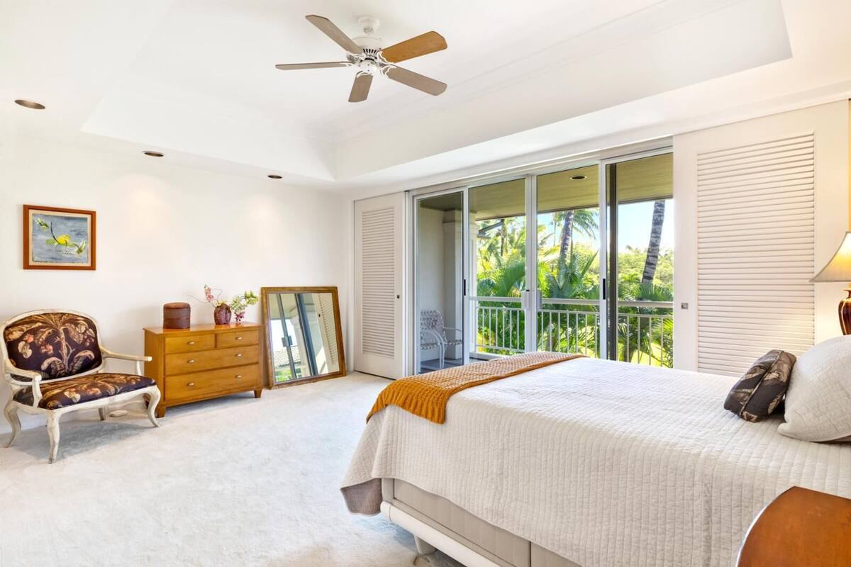 Un dormitorio con cama, cómoda, silla, ventilador de techo y balcón con vista a palmeras.
