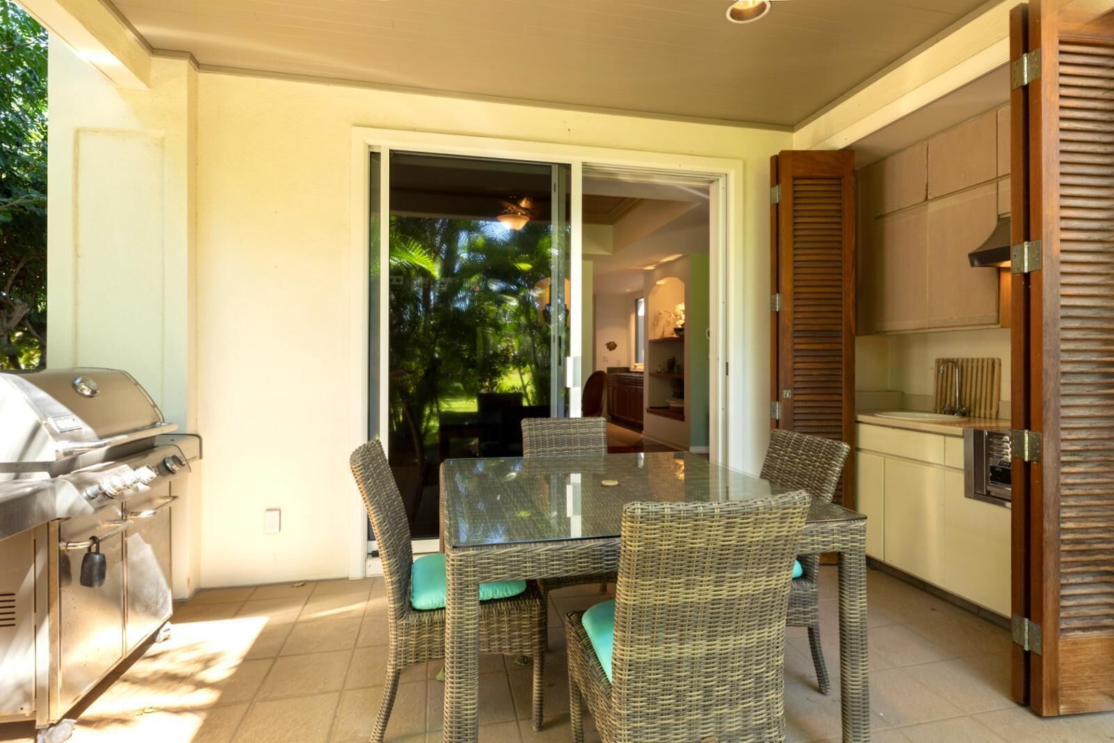 An outdoor dining area with wicker furniture, a grill, and a view of a tropical garden.