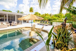 Un espace piscine agrémenté de chaises longues, d’un parasol en chaume et de plantes tropicales.