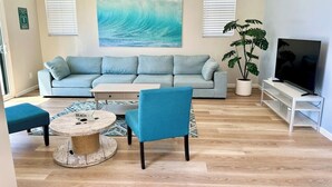 A living room with a sofa, a coffee table, a chair, a TV stand, and a large painting of a wave. - Coastal Compound - Two Fully Equipped 3BRs - 31+ Day Stay (Oceanside)