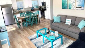 A modern kitchen with white cabinets, a dining area with teal chairs, a grey sofa with teal cushions, and a glass coffee table with a bottle and a menu. - Coastal Compound - Two Fully Equipped 3BRs - 31+ Day Stay (Oceanside)