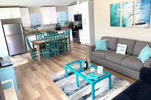 A modern kitchen with white cabinets, a dining area with teal chairs, a grey sofa with teal cushions, and a glass coffee table with a bottle and a menu.