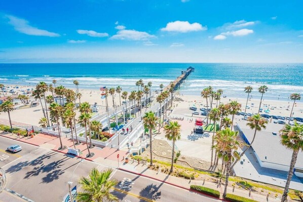 A coastal cityscape with a beach, palm trees, and a pier.