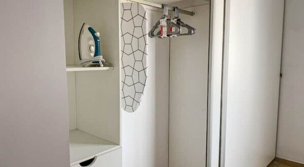 A white closet with a hanging rack, a shelf with an iron, and a geometric-patterned shower curtain.