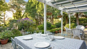 Un área de comedor al aire libre con una pérgola cubierta, una mesa preparada para cuatro personas con platos, vasos y cubiertos, rodeada de exuberante vegetación y plantas en flor.