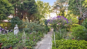 Un sendero en un jardín con una estatua, abundante vegetación y plantas en flor.