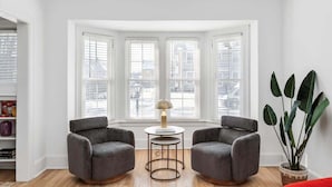 A room with a bay window, two grey armchairs, a small round table with a lamp, and a large potted plant. - Sleeps 14! 10 Mins to Downtown + Driveway Parking (Detroit)