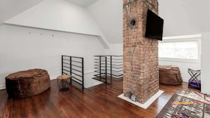 A loft-style room with a sloped ceiling, exposed brick wall, and a mounted TV. - Sleeps 14! 10 Mins to Downtown + Driveway Parking (Detroit)