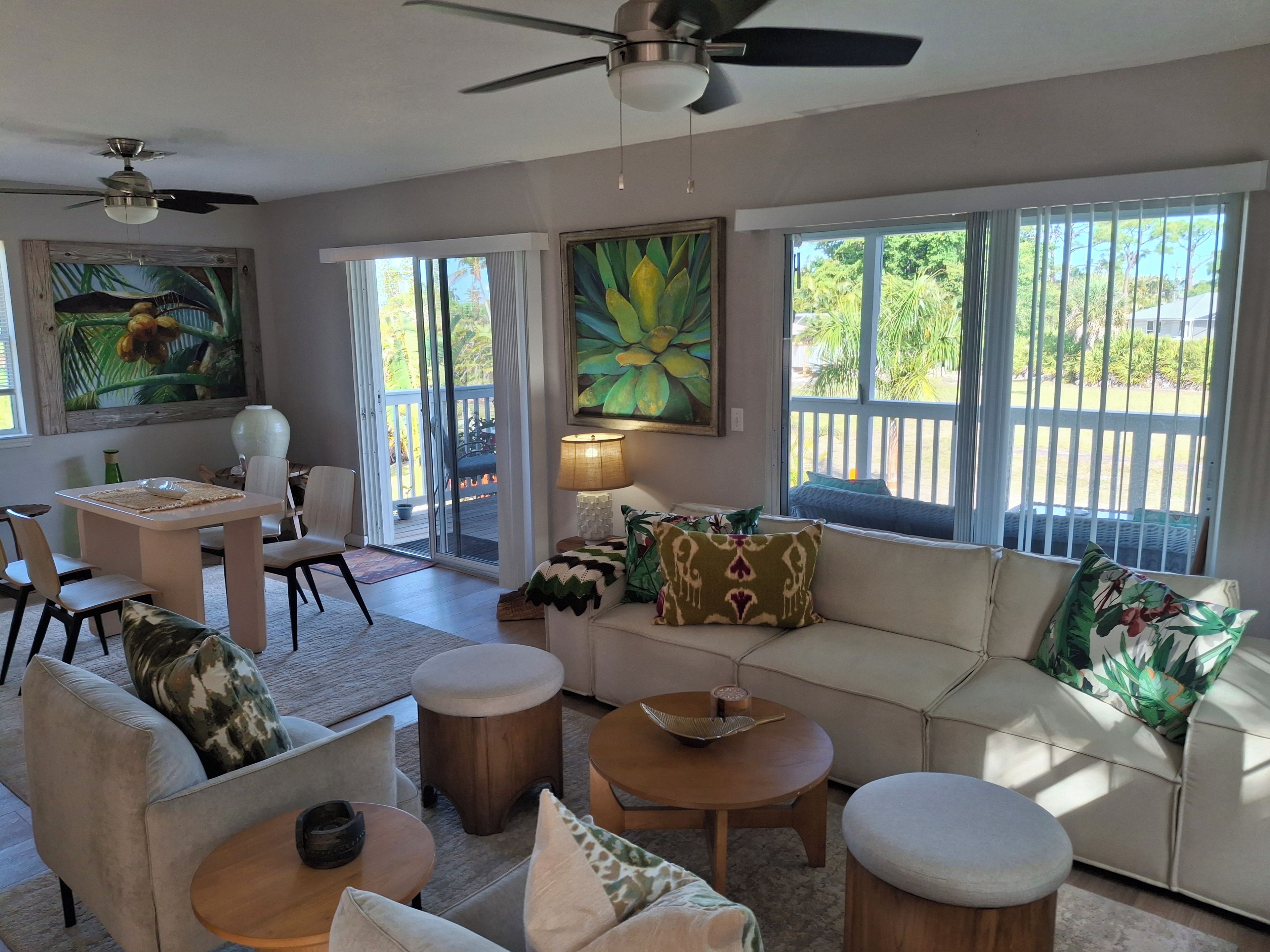 A spacious living area with a sofa, chairs, a coffee table, and a dining table. There are large windows with a view of greenery outside.