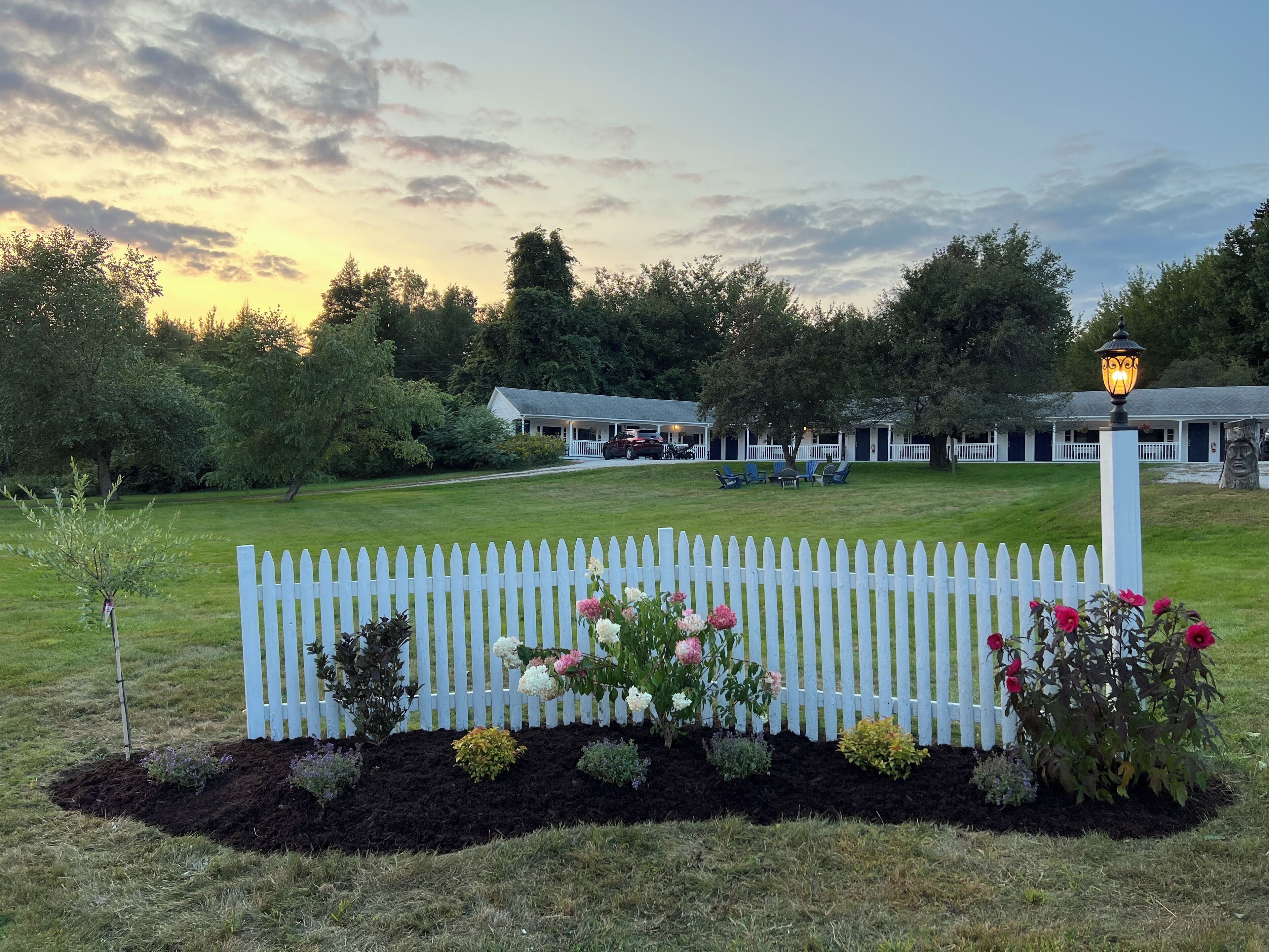 A white picket fence with a garden bed in front of a building with a porch and a lamp post.