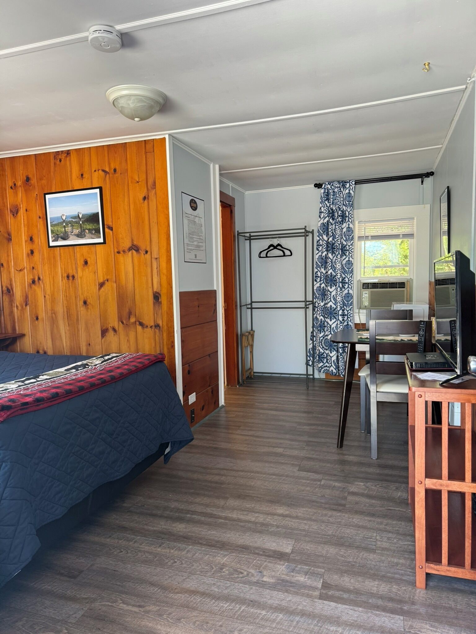 A room with a bed, a desk, a chair, a TV, and a window with patterned curtains.