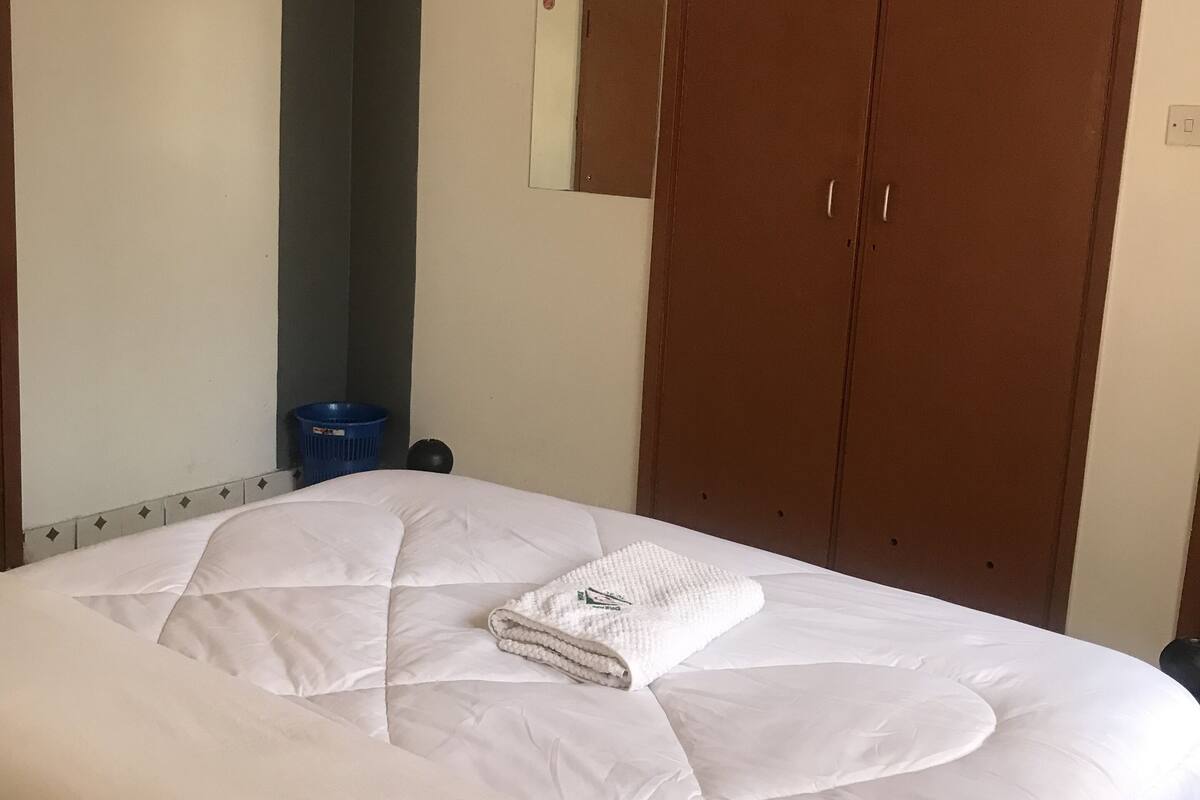 Una cama bien hecha con una colcha blanca y una toalla doblada, un armario de madera y un espejo en la pared.