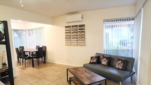 A living room with a sofa, a coffee table, a dining area with chairs, and a large window with blinds. - SALE! Peaceful, Clean, 4x Queen Bed, Spacious! (Hemet)