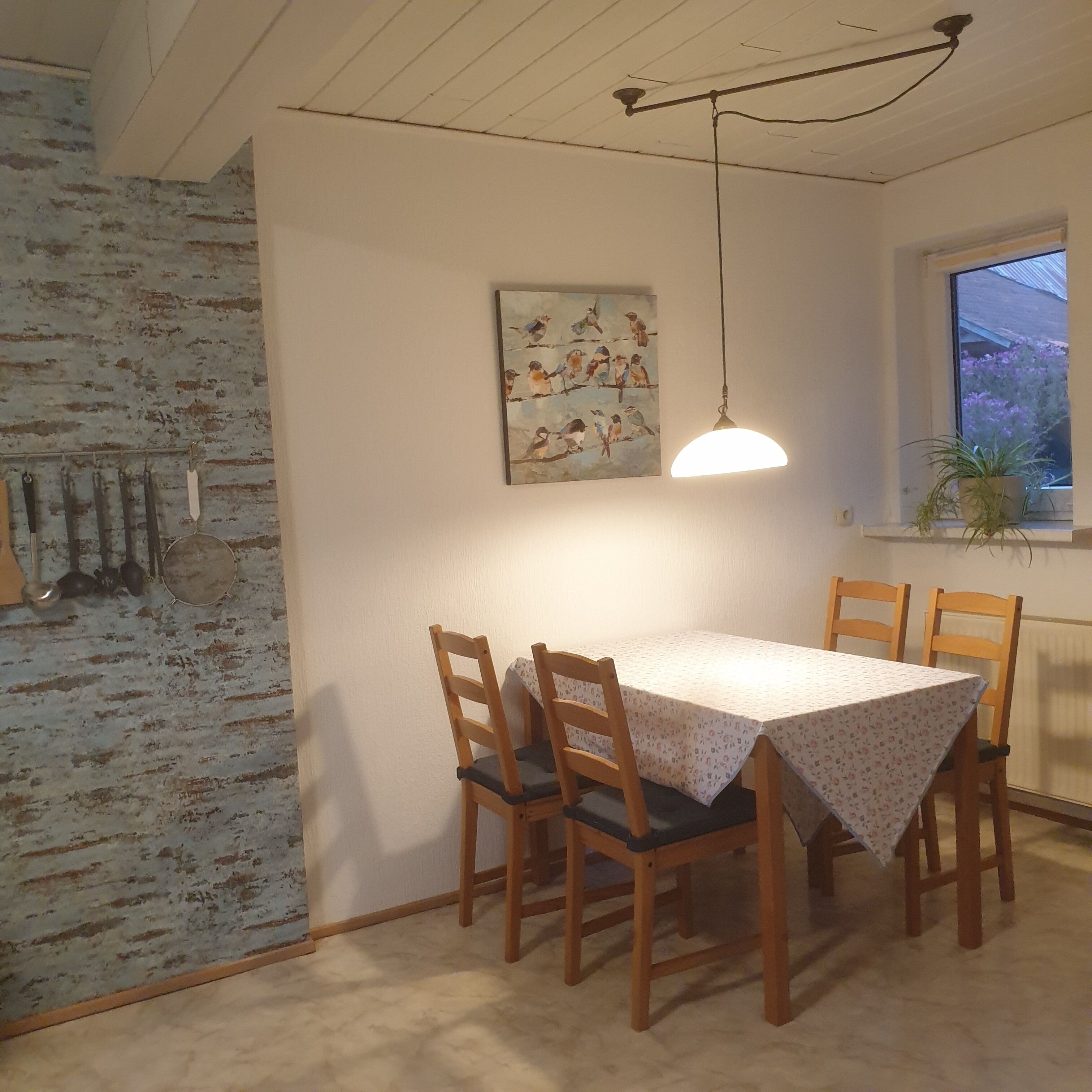 Un comedor con una mesa y sillas, una lámpara colgante, un cuadro en la pared con pájaros y una ventana con vistas al verdor.