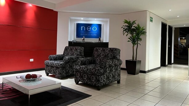 A modern business hotel lobby with a red accent wall, two patterned sofas, a reception desk, and a potted plant. - NEO BUSINESS HOTEL (Culiacán)