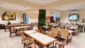 A spacious dining area with wooden tables and chairs, a central green pillar, and a kitchen in the background. - NEO BUSINESS HOTEL (Culiacán)