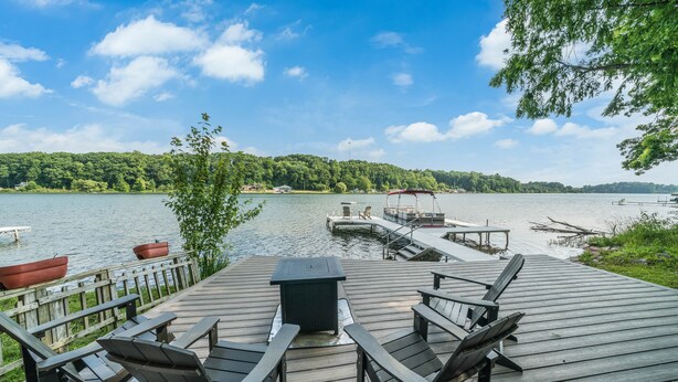 Une terrasse en bois avec des fauteuils Adirondack, une table et un foyer, le tout offrant une vue sur un lac où l’on peut apercevoir des bateaux et un quai.