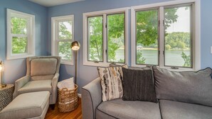 Une pièce avec un canapé gris, un fauteuil gris et un lampadaire. De grandes fenêtres offrent une vue sur les arbres et l’eau.