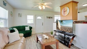 A living room with a green sofa, a wooden coffee table, a ceiling fan, a clock, and a TV displaying a beach scene. - Charming & Cozy: Close to Beach & Shops ~ Parking! (Port Charlotte)