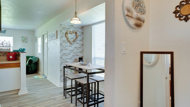 A modern kitchen with a brick accent wall, a dining area with a white table and black chairs, and a hanging light fixture. - Charming & Cozy: Close to Beach & Shops ~ Parking! (Port Charlotte)