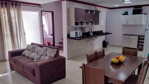 A living room with a sofa and a dining table with chairs, a kitchen area with cabinets and appliances, and a view through an open door.