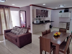 A living room with a sofa and a dining table with chairs, a kitchen area with cabinets and appliances, and a view through an open door.