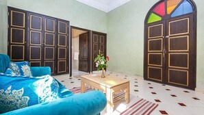 A room with a blue sofa, a wooden coffee table with a vase of flowers, and large wooden doors with a colorful window above. - Hotel Riad Qodwa  (Marrakesh)