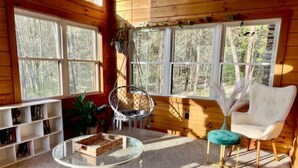 Un interior cálido de cabaña con una mesa de centro de vidrio, dos sillones, una estantería y ventanas amplias que dan a un bosque.
